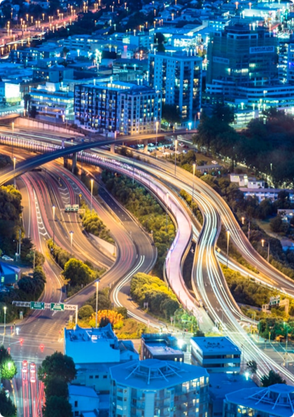 Aerial night view of a city with a multi-level highway interchange, where long-exposure light trails curve between buildings and trees.